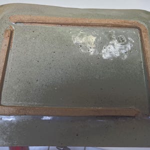 May include: A rectangular ceramic tray with a glossy, speckled sage green glaze. The underside features a raised, unglazed terracotta-colored rectangular foot rim. A small, stamped letter B is visible on the glazed surface near the corner.