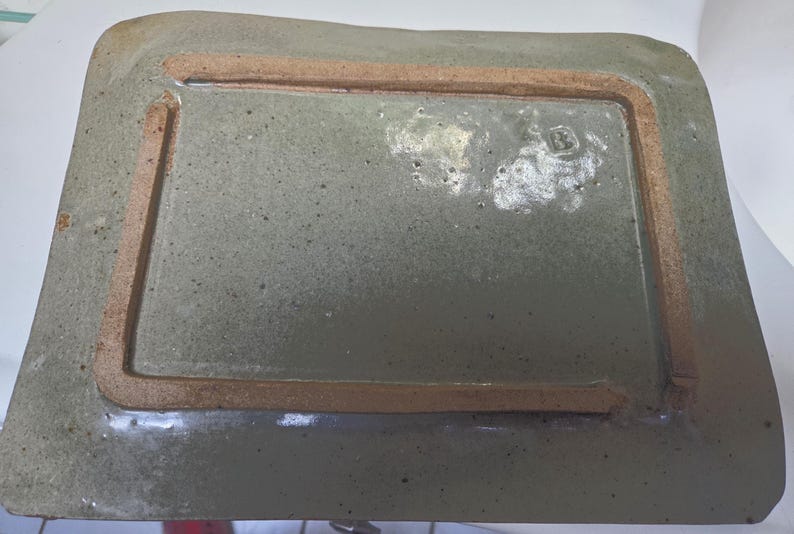 May include: A rectangular ceramic tray with a glossy, speckled sage green glaze. The underside features a raised, unglazed terracotta-colored rectangular foot rim. A small, stamped letter B is visible on the glazed surface near the corner.