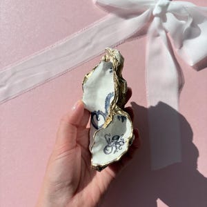 May include: A decorative oyster shell dish featuring a white interior with a detailed blue octopus illustration and gold-painted edges. The shell has a natural, irregular shape and is presented as a small, functional trinket tray for storage.