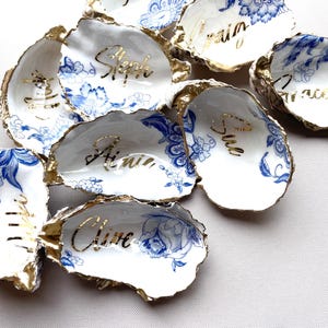 Può includere: Una collezione di conchiglie di ostrica bianche con bordi dorati e disegni floreali blu. Ogni conchiglia ha un nome scritto in oro. Le conchiglie sono disposte su una superficie bianca.