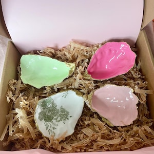 Op de afbeelding: Vier decoratieve oesterschelpen in een doos. De schelpen zijn geverfd in lichtgroen, roze en lichtroze, met gouden randen. Eén schelp heeft een witte basis met een groen bloemmotief. De schelpen zijn genesteld in bruin verpakkingsmateriaal.