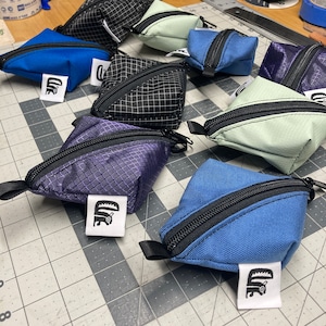 May include: A collection of eight small, triangular zippered pouches in various colors, including blue, green, black, and purple. Each pouch has a black zipper and a small white label with a black graphic.