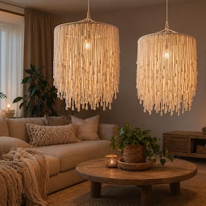 May include: Two cream-colored macrame pendant lights hang in a living room. The lights have a cylindrical shape with long, dangling strands. The room features a beige sofa, a wooden coffee table, and a potted plant, creating a cozy, bohemian atmosphere.