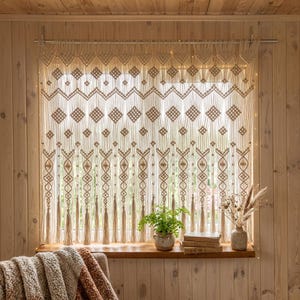 Könnte beinhalten: Beiger Makramee-Fenstervorhang mit geometrischem Muster. Der Vorhang hat Rauten und Quasten. Eine Fensterbank aus Holz beherbergt eine kleine Topfpflanze, Bücher und eine Vase mit getrockneten Blumen.