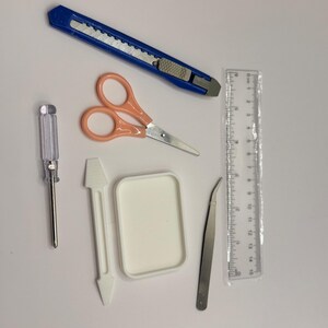 May include: An assortment of craft tools including a blue utility knife, peach-colored scissors, a clear ruler marked in inches, a small screwdriver, a white plastic scraper, a white container, and tweezers.
