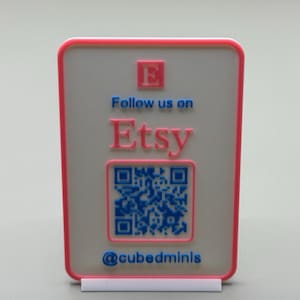 May include: A rectangular sign with a pink border and a white background. The sign features the text "Follow us on Etsy" in blue and pink, along with a QR code and the username "@cubedminis". The sign is displayed on a white stand.
