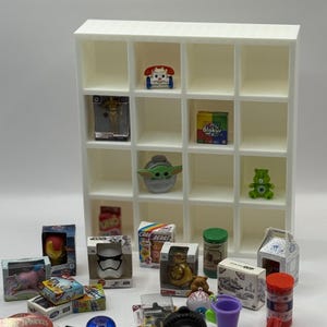 May include: A white miniature display shelf filled with various small toys and collectibles. Items include a Baby Yoda figure, a green Care Bear, and miniature boxed toys. The shelf is empty in some compartments, and the toys are arranged on a white surface.