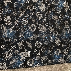 May include: Black fabric with a white space-themed pattern. The pattern includes rockets, planets, stars, aliens, and spaceships.