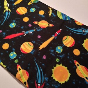 May include: Black fabric with a repeating pattern of planets, stars, and rockets in shades of blue, green, yellow, orange, and red.