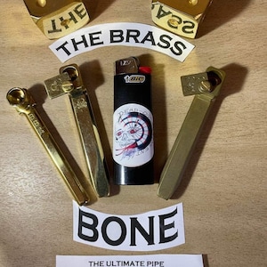 May include: Three gold metal pipes with a skull and crossbones design, a lighter, and two gold dice with the words "THE BRASS" and "BONE" on them. The dice are on a wooden surface.