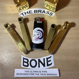 May include: Four gold-colored metal pipes, two gold dice, and a black lighter with a skull and dartboard graphic. The dice read "23" and "THE". The pipes are arranged in a semi-circle around the lighter. The text "THE BRASS" and "BONE" are visible. The text "THE ULTIMATE PIPE DESIGNED FOR THE TRUE SMOKER" is also visible.