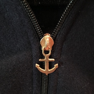 May include: Close-up of a navy blue jacket zipper with a gold-colored anchor-shaped pull. The zipper is black, and the fabric has a soft texture. The anchor pull adds a nautical touch to the garment.