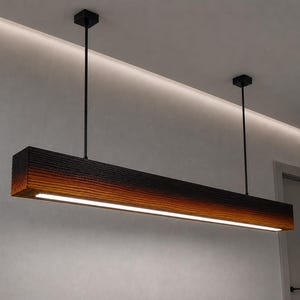 May include: A linear pendant light fitting featuring a long, rectangular wooden beam with a charred, gradient finish transitioning from black at the top to a warm orange-brown at the bottom, suspended by two thin black rods from the ceiling.