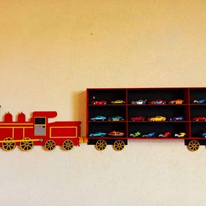 Könnte beinhalten: Ein rotes Holzregal in Form einer Lokomotive mit gelben und grauen Akzenten. Die Regalwagen zeigen eine Sammlung von Spielzeugautos in verschiedenen Farben. Das Regal ist an einer beigen Wand befestigt.