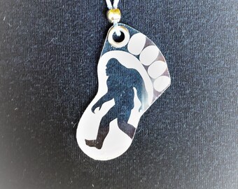 May include: A silver-colored pendant necklace featuring a Bigfoot silhouette within a footprint design. The pendant is suspended from a white cord with a small gold bead. The background is a dark, textured surface.