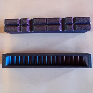 May include: Two rectangular, 3D-printed objects. The top one is dark purple with a textured surface and a series of rectangular cutouts. The bottom object is black with a series of vertical blue dividers inside. Both objects are on a light brown surface.