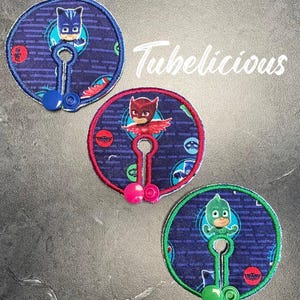 Puede incluir: Tres parches de tela circulares con personajes de dibujos animados de PJ Masks. Cada parche tiene un personaje diferente, con un diseño de ojo de cerradura y un cierre de botón. Los parches son azules, rojos y verdes, con el texto "Tubelicious" en blanco.