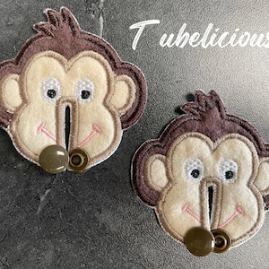 May include: Two monkey-shaped fabric appliques. Each applique features a light brown face, dark brown accents, and a button closure. The text "Tubelicious" is in white script in the upper right corner.