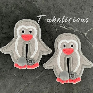 May include: Two gray penguin-shaped fabric appliques with white accents, red beaks and feet, and gray snap closures. The word "Tubelicious" is written in cursive above the penguins.