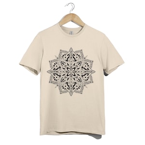 Puede incluir: Una camiseta color crema con un diseño de mandala negro detallado. El mandala es un patrón circular simétrico con intrincados elementos florales y geométricos. La camiseta está colgada en una percha de madera.