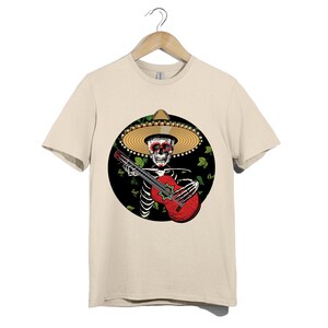 May include: Beige t-shirt featuring a skeleton playing a red guitar, wearing a sombrero. The skeleton is set against a black circle with green leaves. The guitar has a star-shaped sound hole.