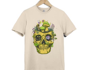 Op de afbeelding: Een beige t-shirt met korte mouwen en een grafische print van een gele schedel waaruit groene paddenstoelen, paarse bloemen en kleine planten groeien aan de bovenkant en uit de oogkassen. Het ontwerp bevat gedetailleerde lijnen en kleuren.