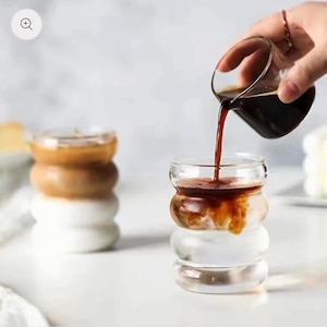 Pode incluir: Canecas de café de vidro transparente com um design arredondado único. Uma caneca é preenchida com uma bebida de café em camadas, enquanto outra está sendo preenchida com café escuro. As canecas estão em uma superfície branca.