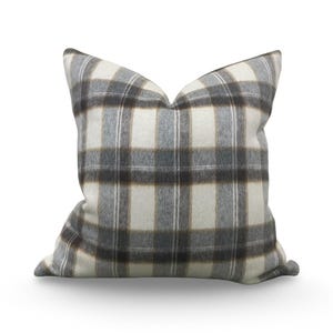 May include: A square throw pillow featuring a plaid pattern in shades of cream, charcoal gray, and soft brown. The fabric has a visible, slightly fuzzy texture, and the design consists of intersecting horizontal and vertical stripes.