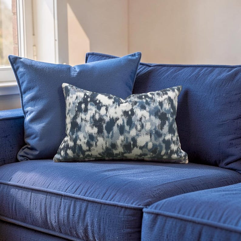 K&ouml;nnte beinhalten: Ein blaues Sofa mit zwei einfarbigen blauen Kissen und einem dekorativen Kissen. Das dekorative Kissen hat ein abstraktes Muster in Wei&szlig;, Blau und Grau. Das Sofa und die Kissen stehen vor einem Fenster.