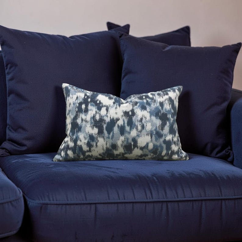 K&ouml;nnte beinhalten: Ein marineblaues Sofa mit mehreren passenden Kissen. Ein Kissen hat ein abstraktes blau-wei&szlig;es Muster. Die anderen Kissen sind einfarbig marineblau. Das Sofa und die Kissen sind aus einem weichen, strukturierten Stoff.