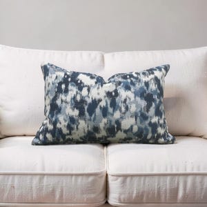 K&ouml;nnte beinhalten: Ein rechteckiges Zierkissen mit einem abstrakten blau-wei&szlig;en Muster. Das Kissen liegt auf einem wei&szlig;en Sofa. Das Muster hat einen Aquarelleffekt, wobei sich Blau- und Wei&szlig;t&ouml;ne vermischen.