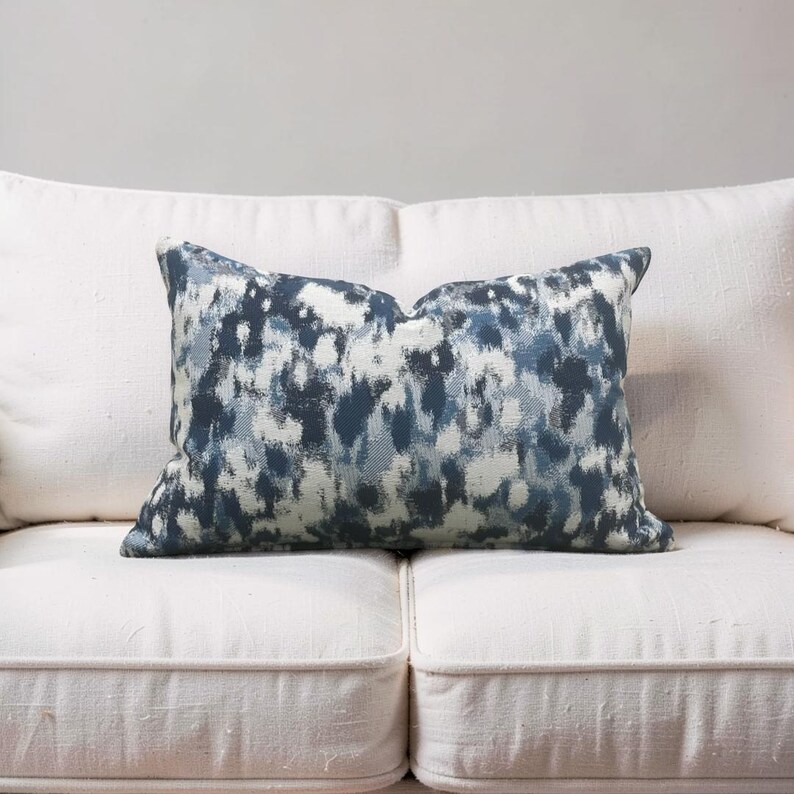 K&ouml;nnte beinhalten: Ein rechteckiges Zierkissen mit einem abstrakten blau-wei&szlig;en Muster. Das Kissen liegt auf einem wei&szlig;en Sofa. Das Muster hat einen Aquarelleffekt, wobei sich Blau- und Wei&szlig;t&ouml;ne vermischen.