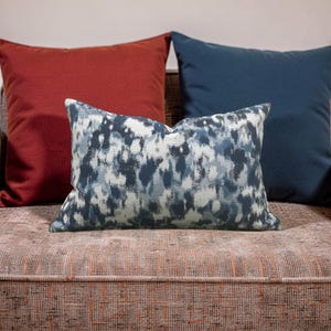 K&ouml;nnte beinhalten: Drei dekorative Kissen auf einem Sofa. Das vordere Kissen hat ein blau-wei&szlig;es abstraktes Design. Dahinter befinden sich zwei einfarbige Kissen, eines in tiefem Rot und das andere in Marineblau. Das Sofa hat einen strukturierten, rotbraunen Stoff.