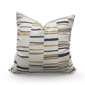 May include: A square throw pillow featuring an off-white base fabric with a repeating pattern of horizontal stripes in shades of mustard yellow, olive green, and charcoal black, arranged in segmented vertical columns across the surface.