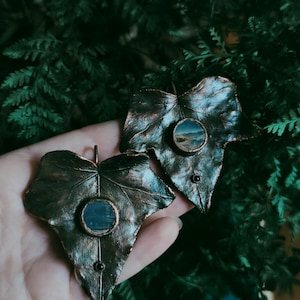 May include: Two bronze-colored leaf-shaped pendants with a round, blue and white image inside each. The leaves have a textured surface and a black gemstone at the bottom.