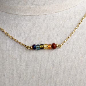 Könnte beinhalten: Eine goldfarbene Halskette mit einem Anhänger in Form eines Balkens. Der Anhänger hat eine Reihe farbiger Perlen in den Farbtönen Lila, Blau, Grün, Gelb, Orange und Rot. Die Halskette wird vor einem neutralen Stoffhintergrund gezeigt.
