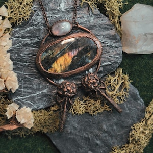 May include: A handcrafted copper pendant necklace featuring a large, oval labradorite stone with iridescent colors. The pendant includes a smaller, oval moonstone and two decorative bird skull charms. The necklace is displayed on a dark gray stone.