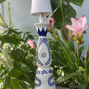 May include: A white ceramic lamp with blue and white floral designs. The lamp is shaped like a tequila bottle and has a white lampshade.