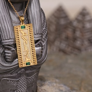 Puede incluir: Collar con colgante rectangular dorado con símbolos jeroglíficos y piedras verdes. El colgante cuelga de una delicada cadena dorada. El collar se muestra sobre un fondo oscuro y texturizado.