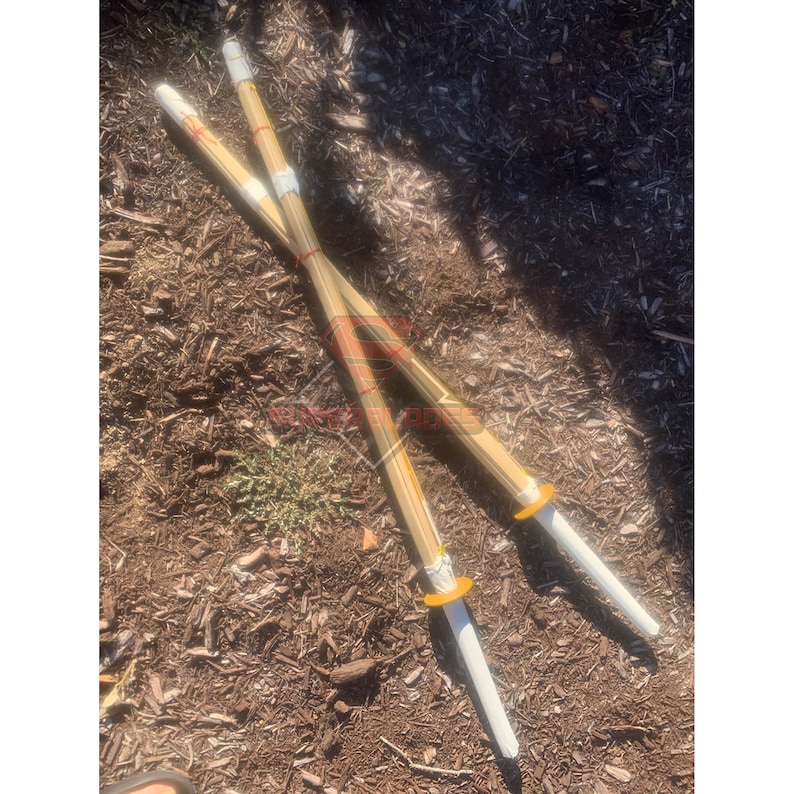 May include: Two wooden practice swords with white tips and yellow accents. The swords are crossed and lying on a bed of brown wood chips.
