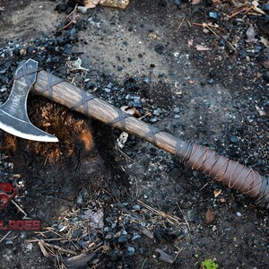 May include: A black axe with a wooden handle wrapped in brown leather. The axe is resting on a tree stump with a dark background.