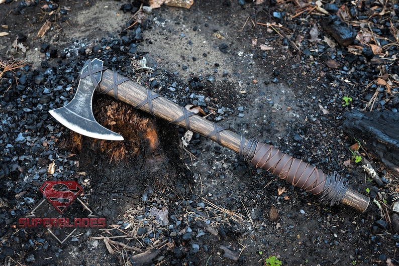 May include: A black axe with a wooden handle wrapped in brown leather. The axe is resting on a tree stump with a dark background.