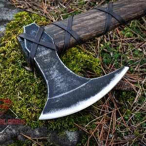 May include: A black axe with a wooden handle wrapped in leather. The axe is lying on a bed of moss and pine needles.