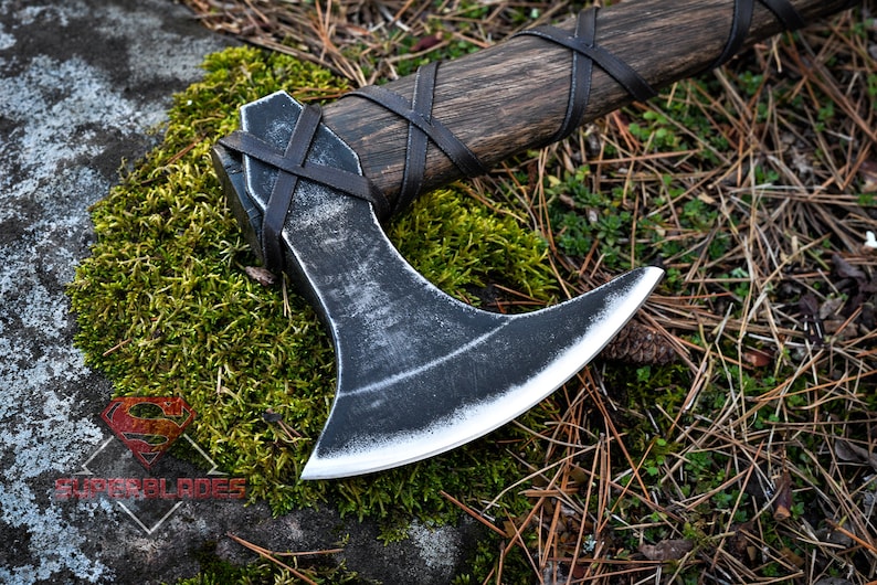 May include: A black axe with a wooden handle wrapped in leather. The axe is lying on a bed of moss and pine needles.