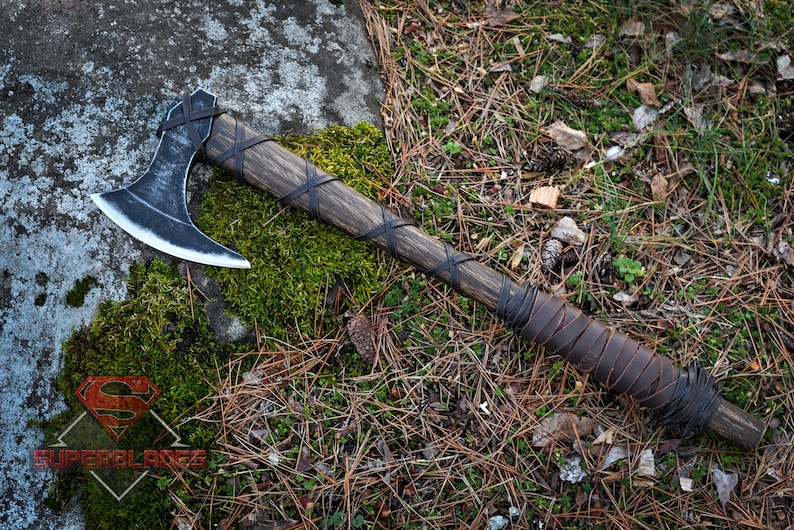 May include: A black and silver Viking axe with a wooden handle wrapped in brown leather. The axe is lying on a bed of pine needles and moss.