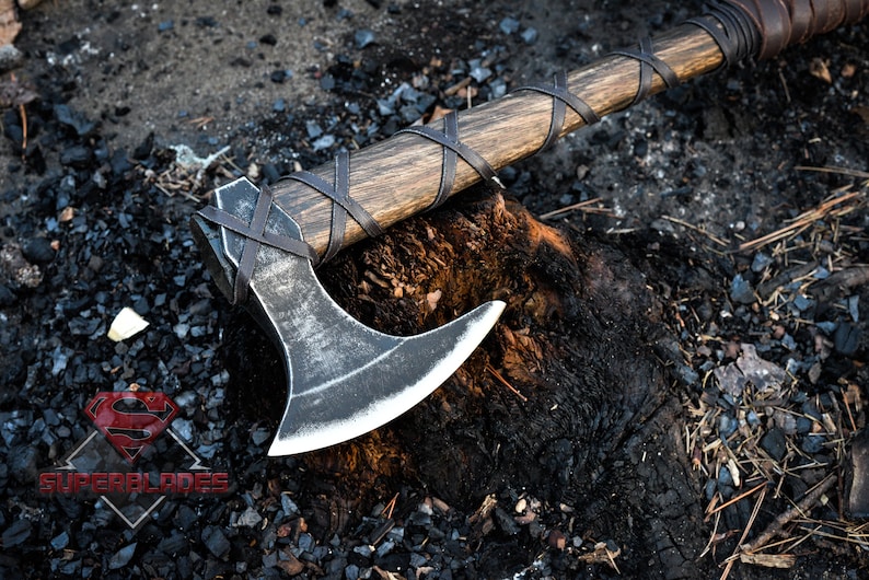 May include: A black axe with a wooden handle wrapped in leather. The axe is resting on a charred tree stump.