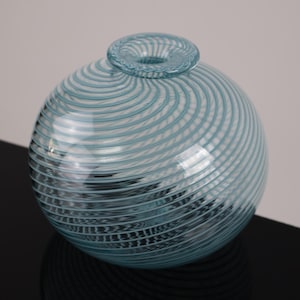 May include: A spherical, light blue glass vase with a small, circular opening at the top. The vase features a spiral design of thin, lighter blue lines that wrap around the body. The vase is sitting on a black surface.