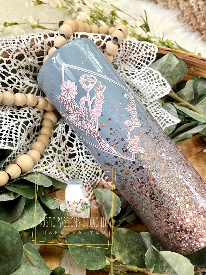 May include: A light blue tumbler with a floral design and the text "get buckled" in rose gold glitter. The tumbler is covered in rose gold glitter.