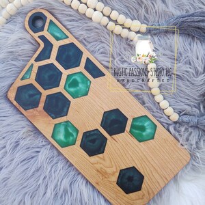 May include: A wooden cutting board with a green and black epoxy resin inlay in a honeycomb pattern. The board has a handle and is handcrafted. The text "Rustic Passions Studio LLC Handcrafted" is visible on the board.