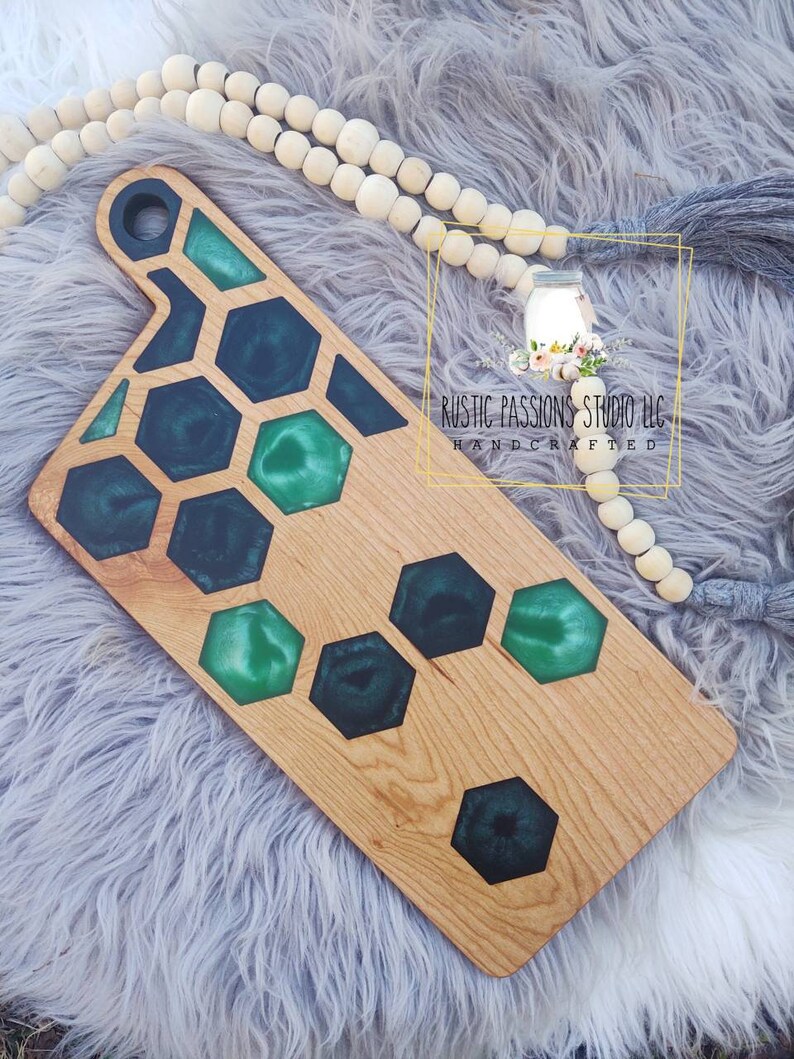 May include: A wooden cutting board with a green and black epoxy resin inlay in a honeycomb pattern. The board has a handle and is handcrafted. The text "Rustic Passions Studio LLC Handcrafted" is visible on the board.